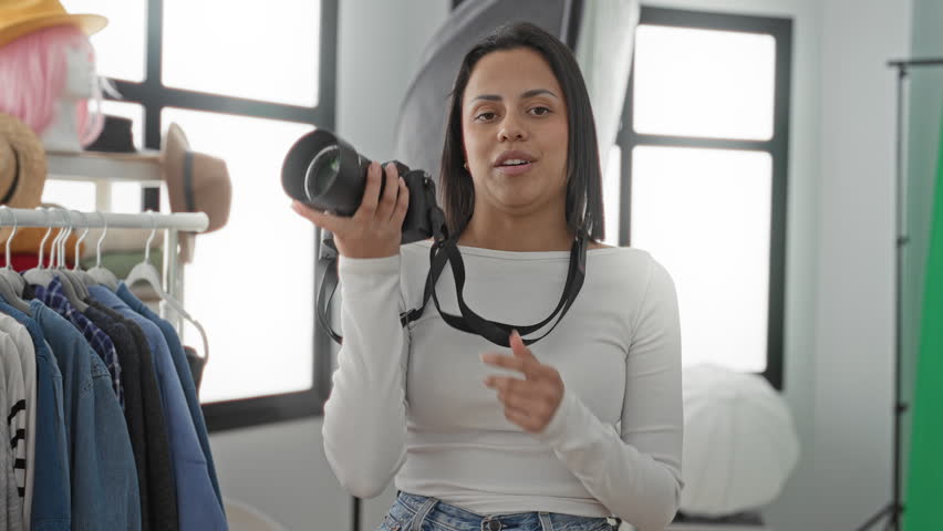 Woman photographer holding dslr camera, speaking to lens and waving hand in a photography studio with clothing rack, hats and softbox visible; confidence tutorial.