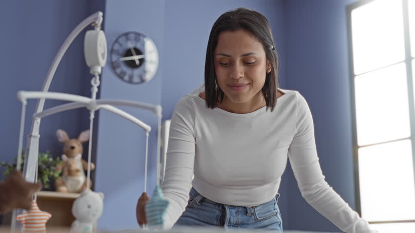 Woman leaning over crib, smiling with hand on rail while adjusting a hanging mobile and plush toys on shelf in a building; maternal tenderness.