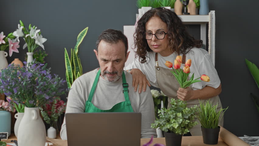 Man florist wearing green apron types on laptop with animated hand gestures while woman florist in beige apron holds orange tulips smiling beside vases in studio; small business joy.