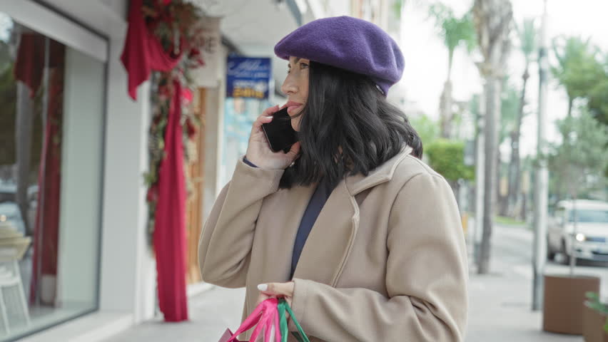 Young hispanic woman holds smartphone to ear and grips colorful shopping bags with one hand while pausing by a storefront on a street; thoughtful.