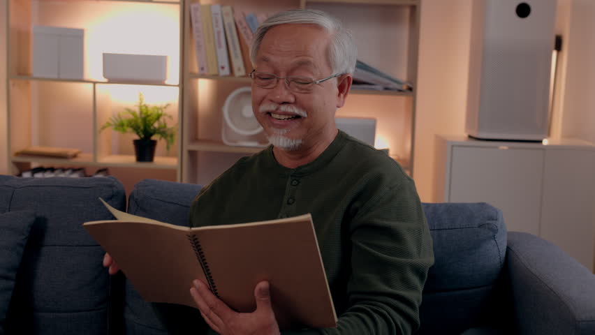 Senior man reading notebook with glasses on sofa in living room at home, elderly Asian senior citizen smiling relaxed, cozy indoor leisure retirement with warm light, casual sweater, beard mustache