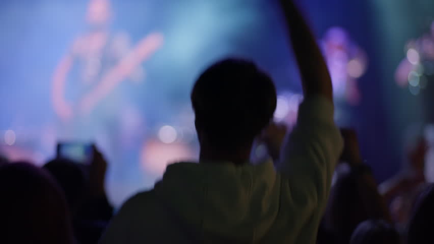 Unrecognizable fan from behind enjoys live music concert. Crowd dances and films with smartphones. Blurry stage with purple lights creates an energetic atmosphere. Shot from audience perspective