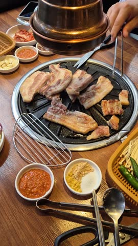 Korean barbecue pork belly grilling on tabletop grill with side dishes.