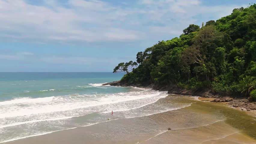 Scenic view of Jeribucaçu Beach in Itacaré, Bahia, Brazil, showing a tranquil tropical coastline with gentle waves, a sandy shore, and a lush forested hillside extending into the ocean under a soft cloudy sky.