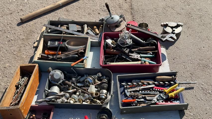 Tools for sale on asphalt showing assorted pliers, wrenches, screwdrivers and sockets in open boxes jars of screws and rusty.