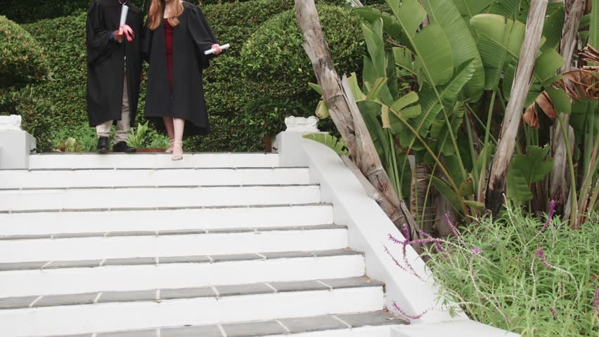 Vertical video: Two grads going down stairs with diplomas, movement causing confetti, graduating. Graduation, diploma, celebration, outdoor, handshake, ceremony, garden