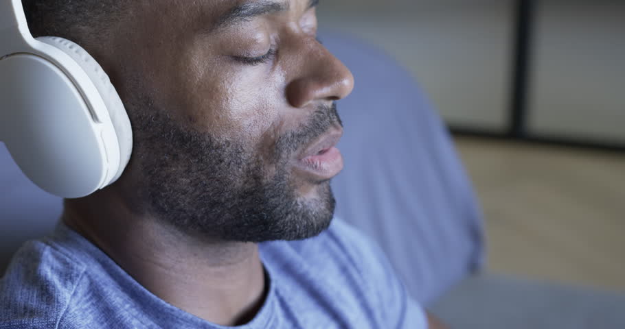 African American man sitting in bedroom wearing headset breathing calming hand on chest, copy space. Headphones, relaxation, mindfulness, adult, calm, home, blue