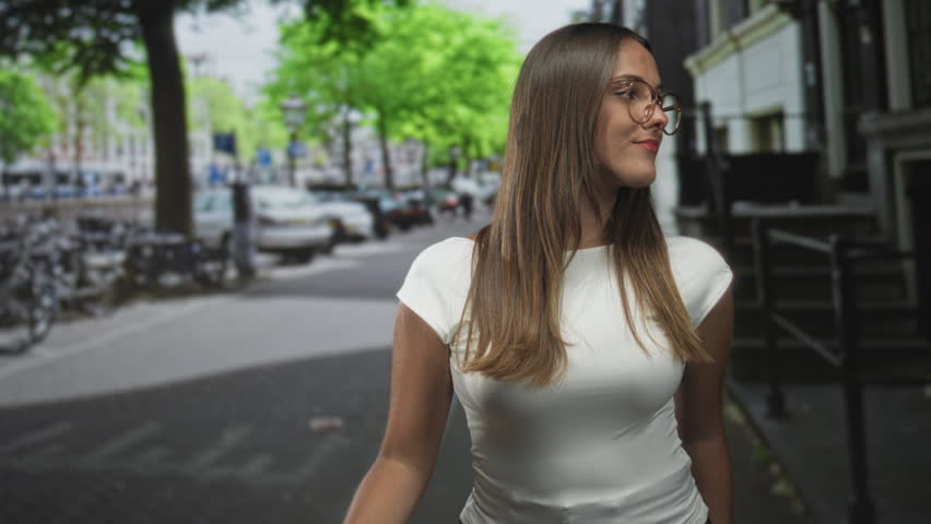 Young woman with glasses looking up while standing on a street beside parked bicycles and a building, wearing a white shirt and long hair; contemplation.