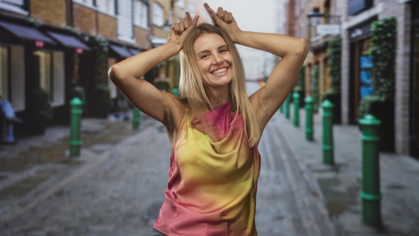 Woman lifts hands to head making horns gesture on a narrow cobblestone street wearing a sleeveless yellow pink top; playful joy.