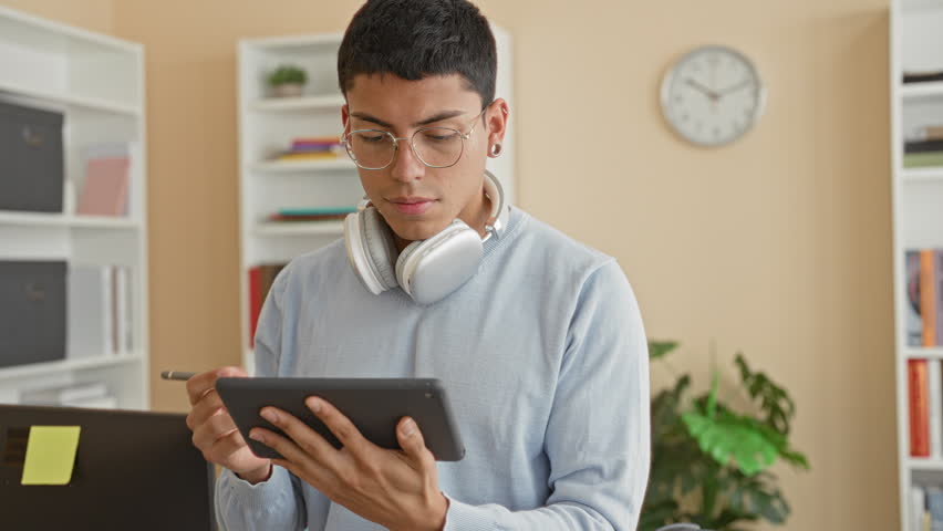 Man holding tablet and stylus, wearing headphones and glasses, tapping screen while standing in a study building; thoughtful focus.