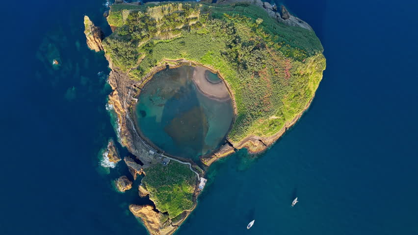 Circular islet lagoon bordered by steep volcanic cliffs rising above Atlantic ocean water in Azores, drone footage