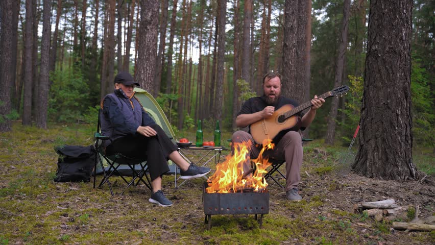 A young couple enjoys a peaceful evening in the woods, where a man plays an acoustic guitar and sings songs to his girlfriend while sitting by a warm, crackling fire near their tent