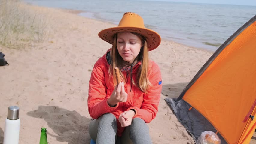 woman eating sausage close-up sitting near tent at lakeside beach campsite, enjoying simple food during outdoor trip, relaxing alone, camping routine and travel lifestyle by water