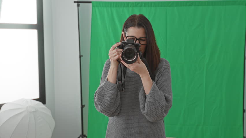 Woman photographer holding camera, hands on lens, smiling in studio with green backdrop; creative joy.