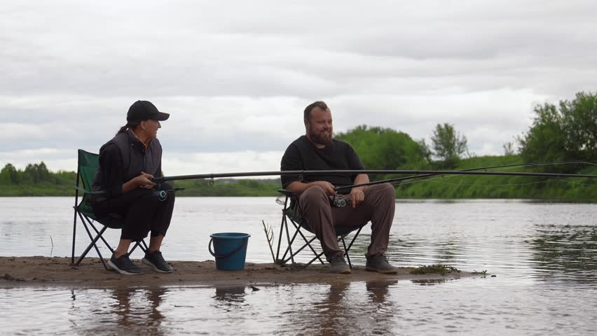 Mature man and woman enjoying a quiet day fishing together on the sandy bank of a river, sitting on folding chairs while patiently waiting for a catch on a peaceful, cloudy day