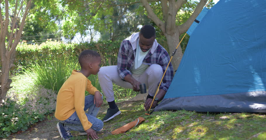 African American father and child arriving near blue tent in grass, hammering stake to secure rope. Shelter, camping, teamwork, family, outdoor, shade, mentorship