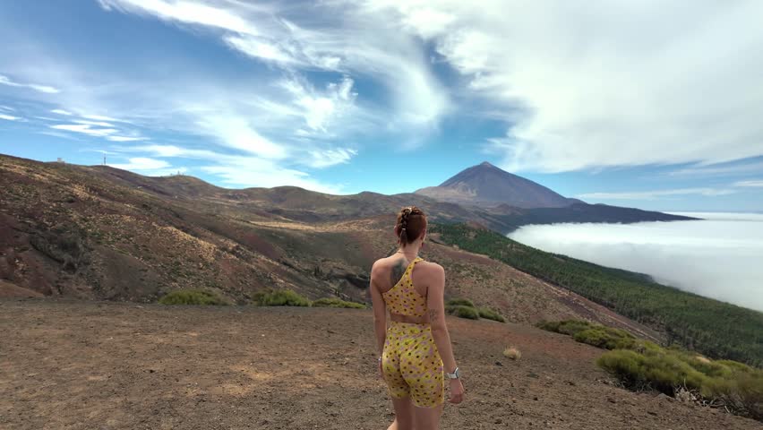 Back view of a young athletic woman reaching a mountain peak, raising her arms in celebration. Scenic view of mount teide above the clouds