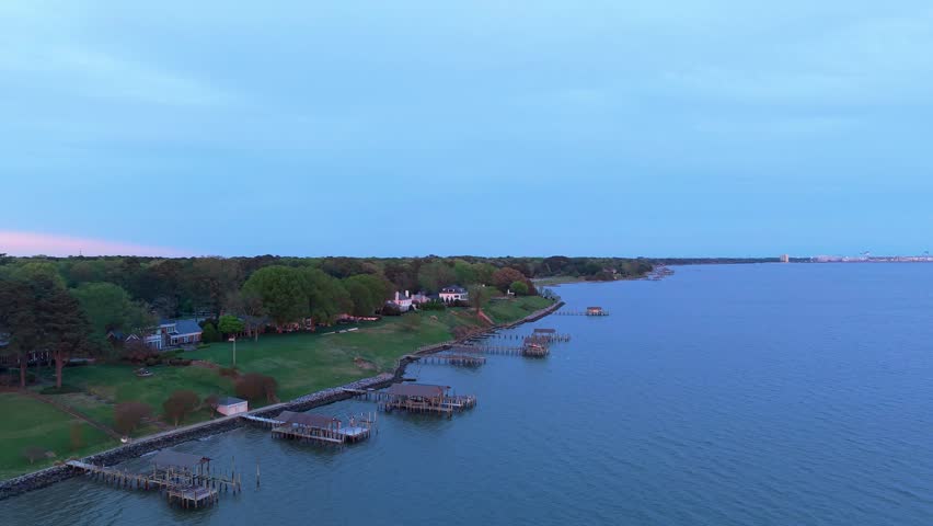 Aerial video of Newport News coastline along the James River, featuring a row of private wooden piers and boat houses along the grassy shoreline at dusk.