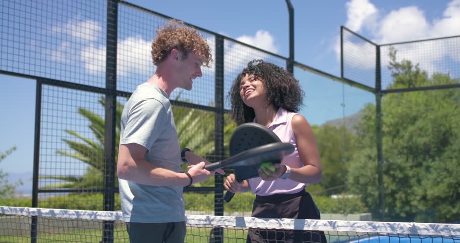 Couple exchanging paddles, woman tossing ball and preparing serve to begin match on paddle court. Duo, joy, laughter, outdoor, sunshine, palm, tropical