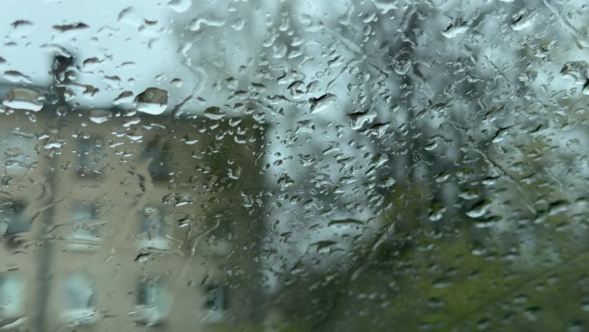 Close-up of water drops on a windowpane. Out-of-focus background reveals a brick apartment building, a street, and green trees in spring.