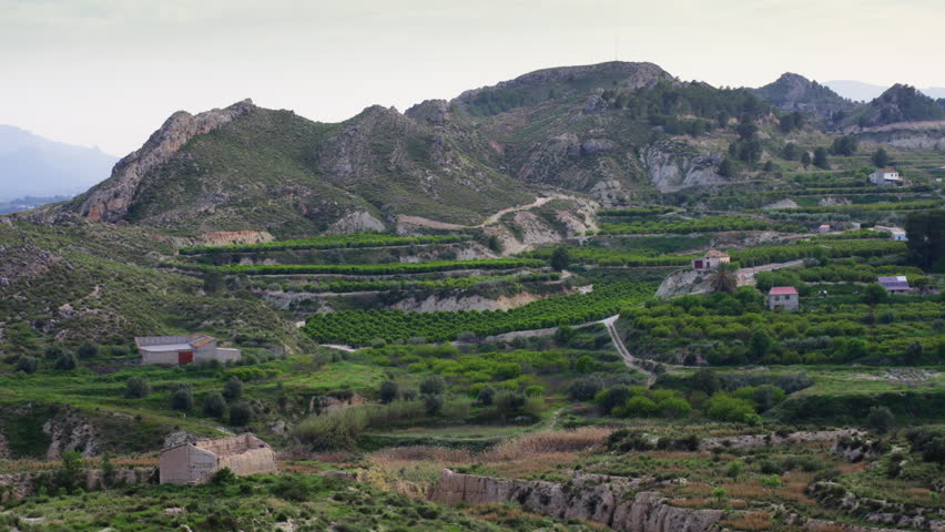 Picturesque rural landscape featuring green agricultural terraces carved into the hillsides of a mountain valley, with scattered farmhouses and an old ruin among fruit tree groves
