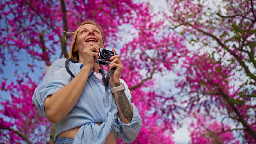 Beautiful smiling woman taking photos with a vintage camera in a park filled with vibrant pink blooming trees. Joyful female photographer enjoying her hobby outdoors during a sunny spring day