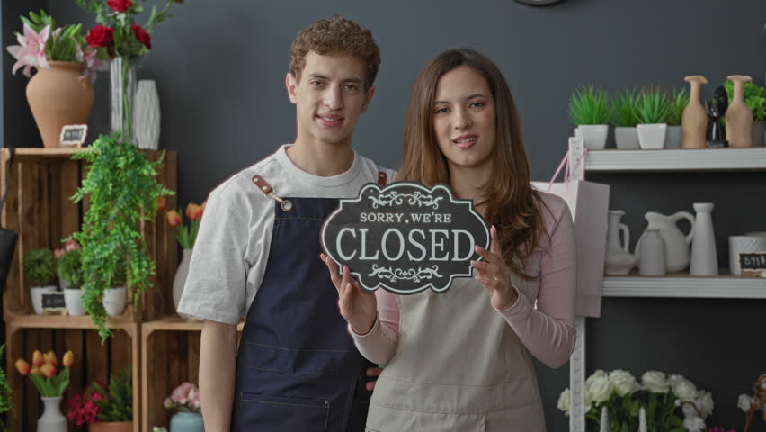 Man and woman hold an open sign together, smiling and pointing to it with visible hands, aprons on inside a shop building; welcoming.
