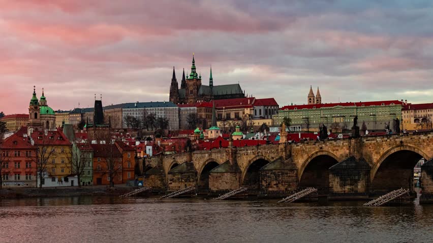Prague. A panoramic view of a cityscape with a castle and a bridge in the foreground. The city is illuminated by the setting or rising sun, casting a golden hue over the buildings and the bridge.