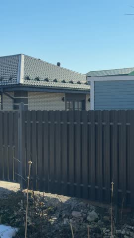 One storey brick cottage behind fence sky sunlit suburban scene with metal slat fence, tiled roof, gravel driveway.