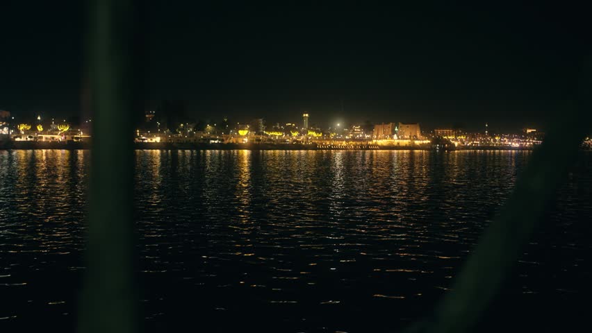 Night view of Luxor Temple ruins from the Nile. Nighttime scene looking across the Nile from the west bank to the east. Luxor Temple ruins are illuminated, glowing against the dark sky. The opposite riverbank is brightly lit, contrasting with the dark water. The scene captures the dramatic nighttime atmosphere of Luxor, highlighting the temple ruins and the river’s reflection under artificial lights.