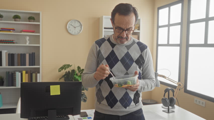 Middle aged man eating salad with fork at computer in office building, holding plastic container by desk and monitor while standing; calm concentration.