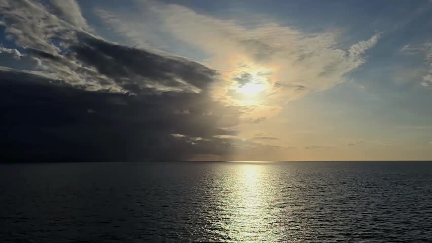 Sun partially obscured by dark clouds over the ocean with golden light reflecting on the water at sunset, creating a dramatic seascape.