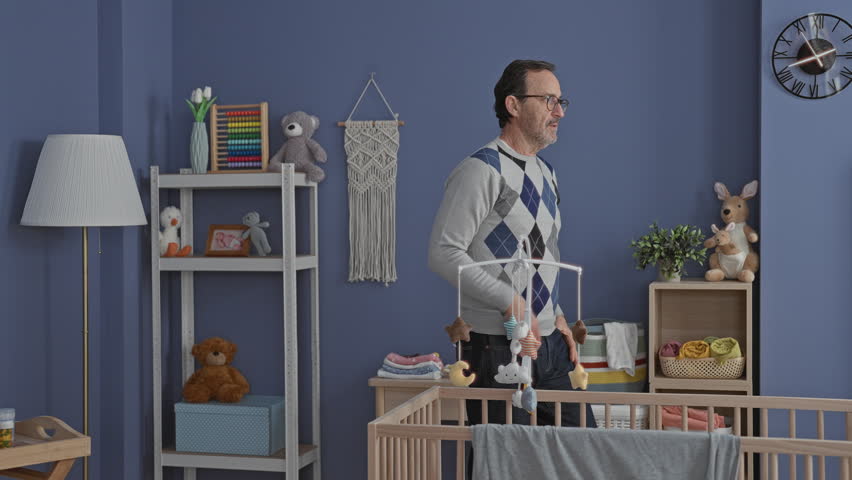 Man with hands on cheeks beside wooden crib and hanging mobile, shelves of toys and clock in nursery building; quiet concern.