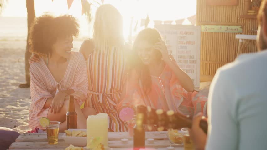 Women trio sitting at beach table, graphic bars sliding over group, man strumming guitar for ads. Outdoor, sand, sunset, friends, casual, acoustic, picnic