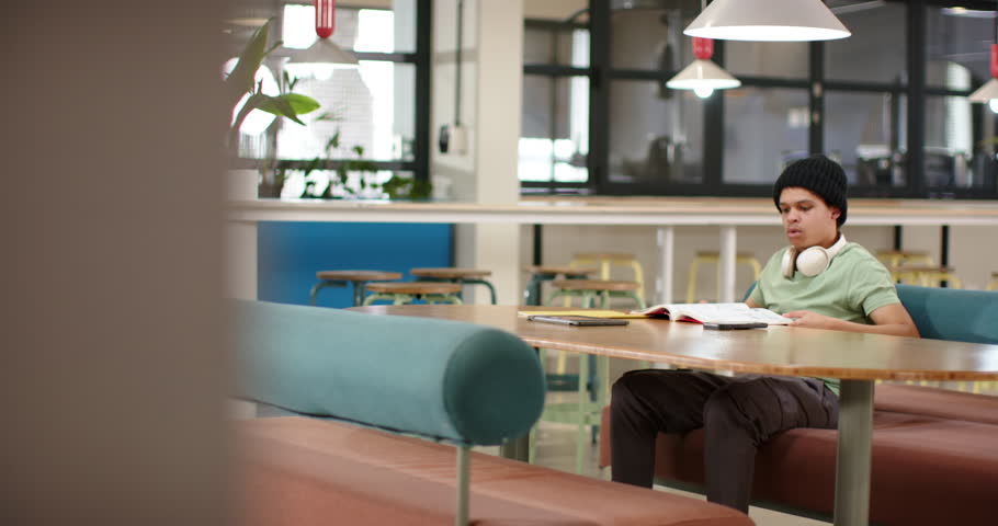 Reaching for red book, African American man turning pages, reading for study at table, copy space. Headphones, beanie, coworking, lounge, tablet, teal, wood