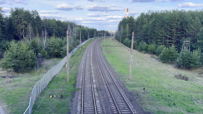 Electric railway tracks winding through a pine forest under a cloudy blue sky, a transport route and a green nature corridor.