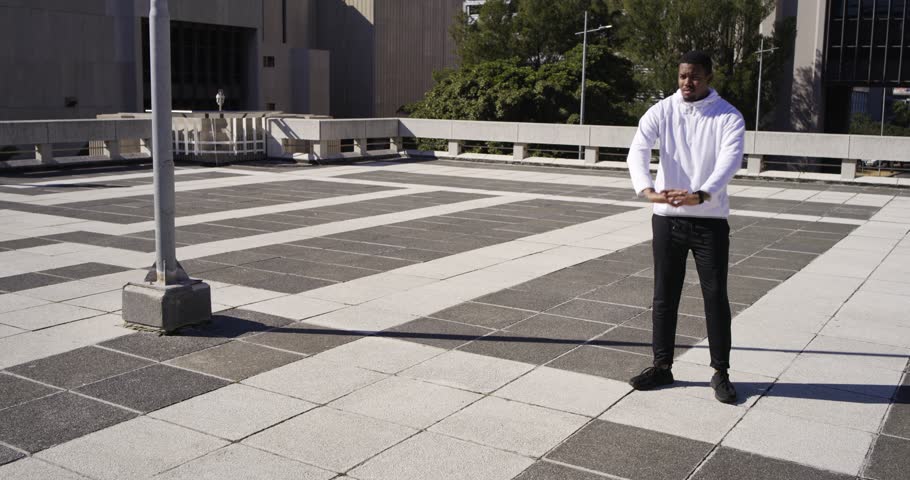 African American man adjusting hoodie, stretching and moving into low lunge on terrace, warming up. Rooftop, outdoor, fitness, movement, urban, daylight, shadows