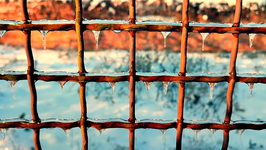 A metal old rusty fence covered in ice on a sunny winter day. Small frozen icicles hang from the metal wire of a fence.