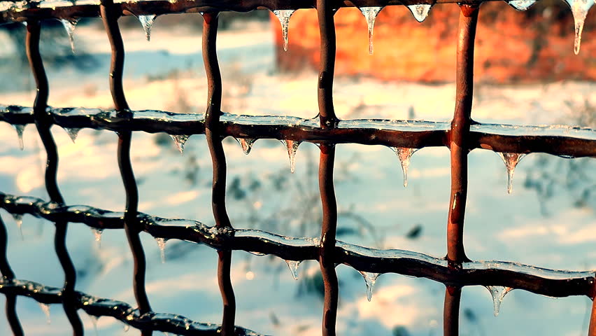 A metal old rusty fence covered in ice on a sunny winter day. Small frozen icicles hang from the metal wire of a fence.