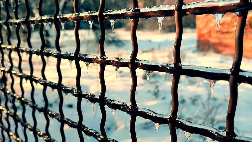 A metal old rusty fence covered in ice on a sunny winter day. Small frozen icicles hang from the metal wire of a fence.