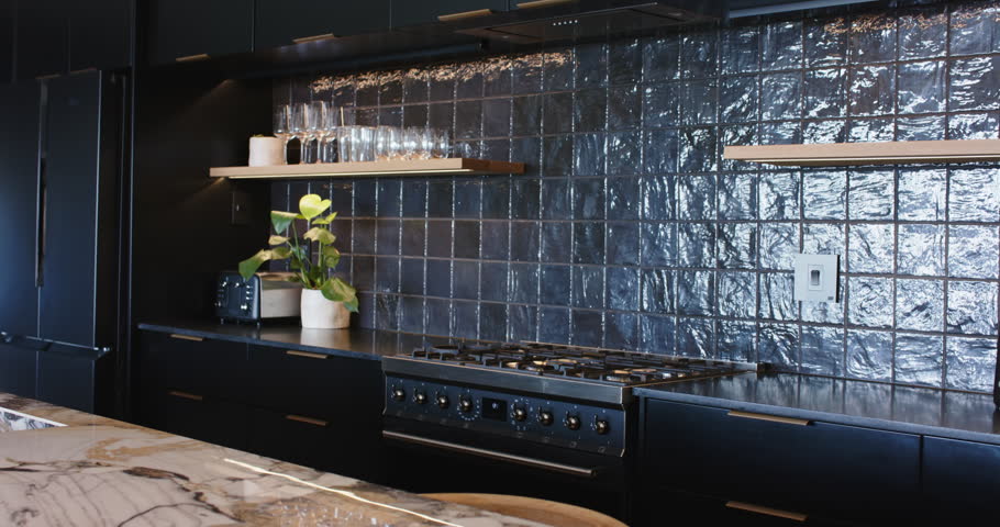 Camera is panning left, revealing black gas range, glossy tiled backsplash, wooden shelf and fridge. Kitchen, modern, dark, glassware, island, marble, monochrome