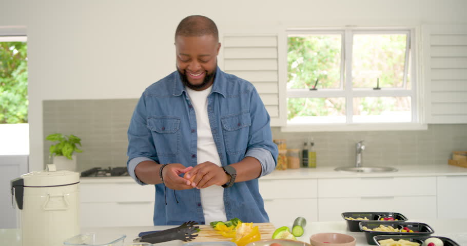 African man in denim holding egg, cracking into cooker and packing meal boxes at kitchen island. Countertop, prep, containers, vegetables, appliance, organization, bright