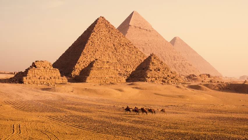 Group of camels and their owners on desert plateau with the Pyramids of Giza in the soft morning light, Egypt. A group of camels with their owners walks across a sandy desert plateau near the Pyramids of Giza at sunrise. The three main pyramids rise in the background, with smaller pyramids nearby. The morning light creates a soft, hazy atmosphere, emphasizing the scale and historic beauty of the site.