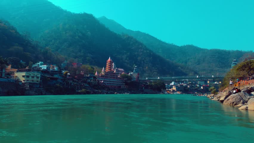 Aerial drone view of Rishikesh showing temple structures, flowing Ganga river, and iconic bridge with surrounding Himalayan landscape.