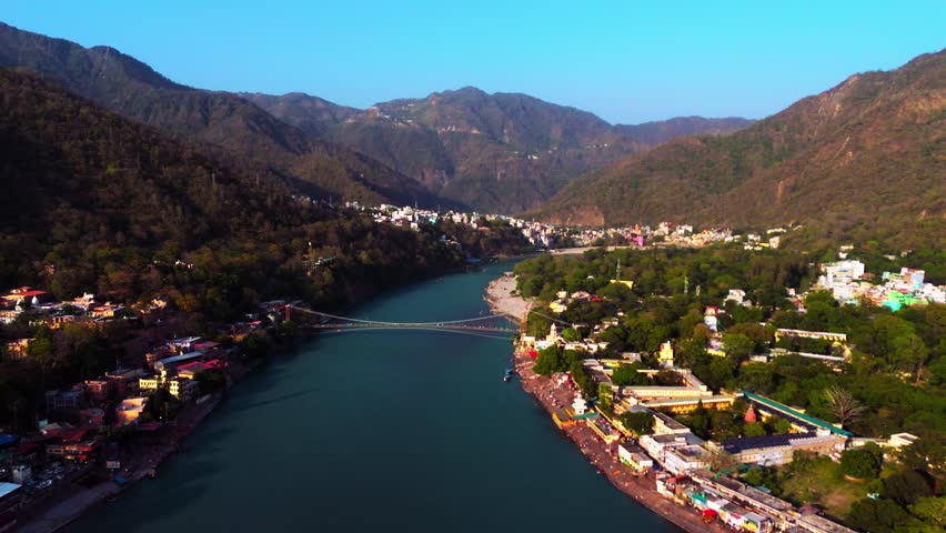 Aerial drone view of Rishikesh featuring the Ganga river, surrounding Himalayan mountains, and iconic suspension bridge (Jhula).