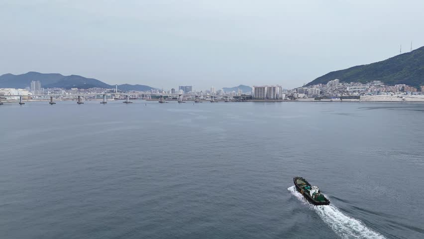 Aerial drone footage of work boat moving across calm sea leaving wake toward Busan city skyline and bridge, minimal seascape with copy space, transport and business concept