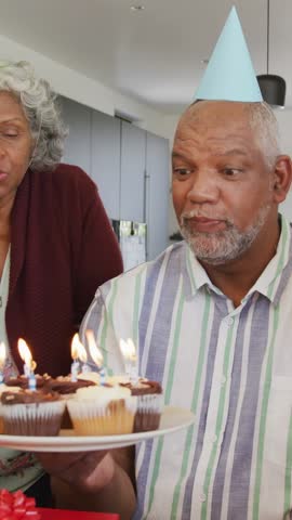 Vertical video: Woman bringing cupcakes, man blowing candles, marking birthday, overlaying bokeh. Celebration, couple, home, cozy, surprise, delight, vertical video