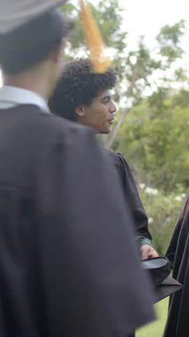 Vertical video: Retrieving cap, diverse graduates in gowns shaking hands, celebrating on lawn. Commencement, achievement, camaraderie, celebration, outdoor, youthful, vertical video