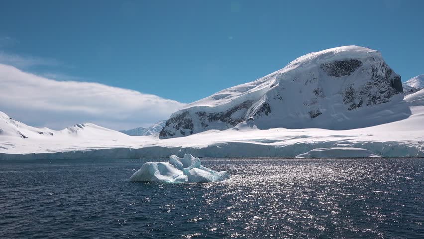Mountains covered with snow stand tall against the blue sky. Icebergs drift in the water, reflecting the landscape. This is a remote area in the Arctic during daytime.