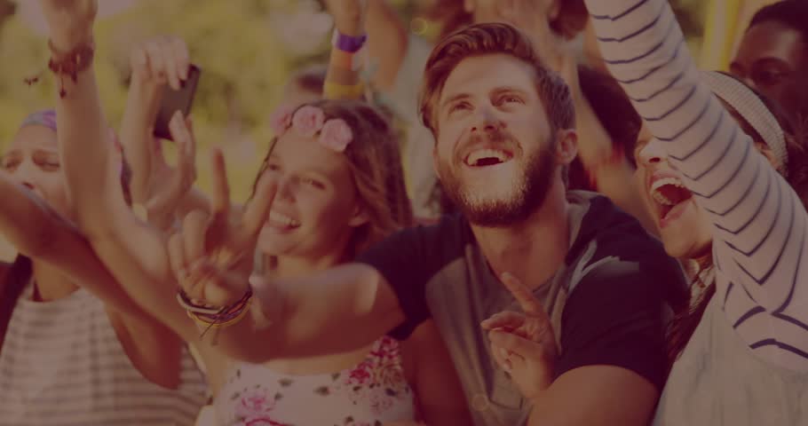 Woman lifting phone starting selfie as bearded man cheering with crowd at fest, gold orbs drifting. Festival, outdoor, frontrow, stage, camera, friends, sunny